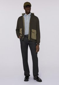 Olivově zelený žebrovaný cardigan s khaki kapsami, přes modrou a bílou pruhovanou košili; spárován s tmavými džíny a černými botami, strukturovaná čepice.