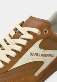 Καφέ και μπεζ αθλητικό παπούτσι Karl Lagerfeld με εκρού κορδόνια και λεπτομέρειες από σουέντ και δέρμα, με το όνομα της μάρκας στην πλαϊνή πλευρά.