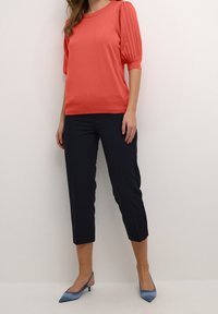 Frau in einer korallenfarbenen Kurzarmbluse, schwarzen Caprilanghosen und blauen spitzen Slingback-Heels, steht vor einem schlichten weißen Hintergrund.