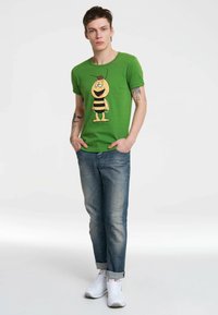 Green cotton T-shirt featuring a cartoon bee graphic in yellow and black. Paired with blue denim jeans and white sneakers.