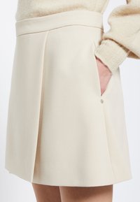 Falda plisada beige con una textura suave y una cinturilla plana. Presenta un bolsillo lateral y un pequeño acento metálico cerca del bolsillo.