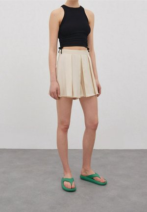 Crop top noir côtelé, mini-jupe plissée beige et sandales tongs vertes portés par un mannequin sur un fond uni.