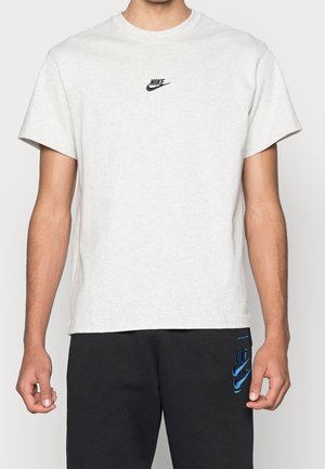 Hombre con camiseta Nike gris claro con logo negro en el pecho y pantalones de chándal Nike negros con logo azul, de pie sobre un fondo liso.