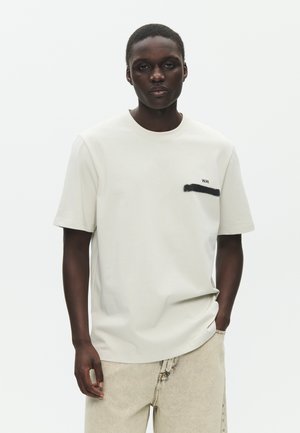 Jeune homme noir portant un T-shirt blanc avec une bande noire et le texte "W.W." sur la poitrine, un pantalon beige, debout devant un fond blanc.