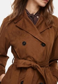 Cappotto trench in suede marrone con design doppiopetto, bottoni neri e una cintura legata in vita. Presenta ampi risvolti e una texture morbida.