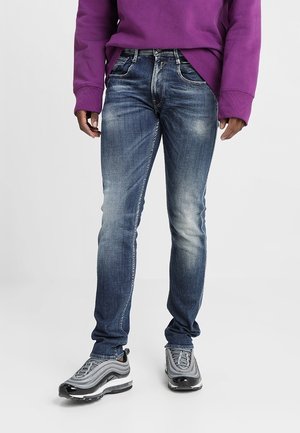 Person trägt ausgewaschene blaue Slim-Fit-Jeans, graue und weiße Turnschuhe sowie ein lila Langarmshirt und steht vor einem einfarbigen hellen Hintergrund.