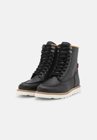 Botas de cuero negro con acabado texturizado, diseño de cordones, suela blanca contrastante y detalles en marrón alrededor de la base.