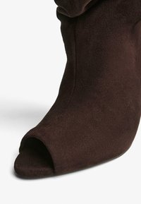 Brown suede ankle boot with an open toe, featuring a sculpted heel and smooth texture. The design includes a slouched upper.