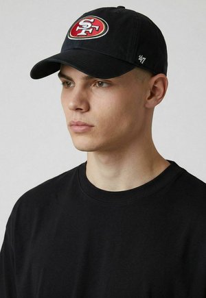 Junger Mann trägt eine schwarze San Francisco 49ers Kappe und ein schwarzes Rundhals-Shirt, schaut leicht nach links vor einem einfarbigen Hintergrund.