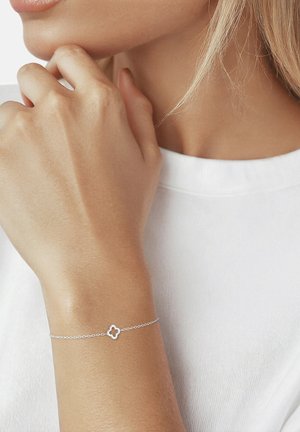 Pulsera delicada de cadena de plata con un pequeño colgante de trébol de cuatro hojas, usada en una muñeca, mano tocando el mentón, camisa blanca visible.