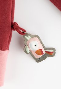 Roter Stoff mit einem pinken Bündchen, das ein abnehmbares, vogelähnliches Etikett in sanften Farben, darunter Pink, Weiß und Orange, mit gestickten Details zeigt.