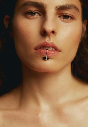 Primer plano del rostro de una mujer con labios brillantes. Un piercing labial plateado presenta un acento triangular negro y una barra vertical. Textura de piel suave.