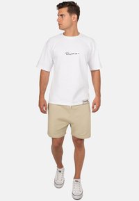 T-shirt bianca a maniche corte con logo nero, abbinata a pantaloni corti beige e sneaker bianche con accenti rossi e lacci.