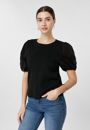 Frau mit dunklen Haaren, schwarzem Oberteil mit transparenten Puffärmeln und blauen Jeans, steht mit einer Hand in der Tasche vor einem einfarbigen Hintergrund.