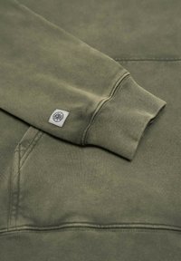 Grön bomullströja med ribbad mudd, en texturerad yta och en liten logotyp på ärmen. Observera den mjuka, dämpade färgen.