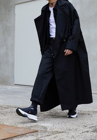 Czarny długi płaszcz na czarnej koszuli i spodniach, w zestawie z czarnymi trampkami i skarpetkami. Płaszcz ma luźny krój i teksturowany materiał.