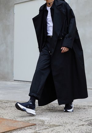 Cappotto lungo nero sopra una maglietta nera e pantaloni neri, abbinato a sneakers nere e calzini. Il cappotto presenta una vestibilità ampia e un tessuto testurizzato.