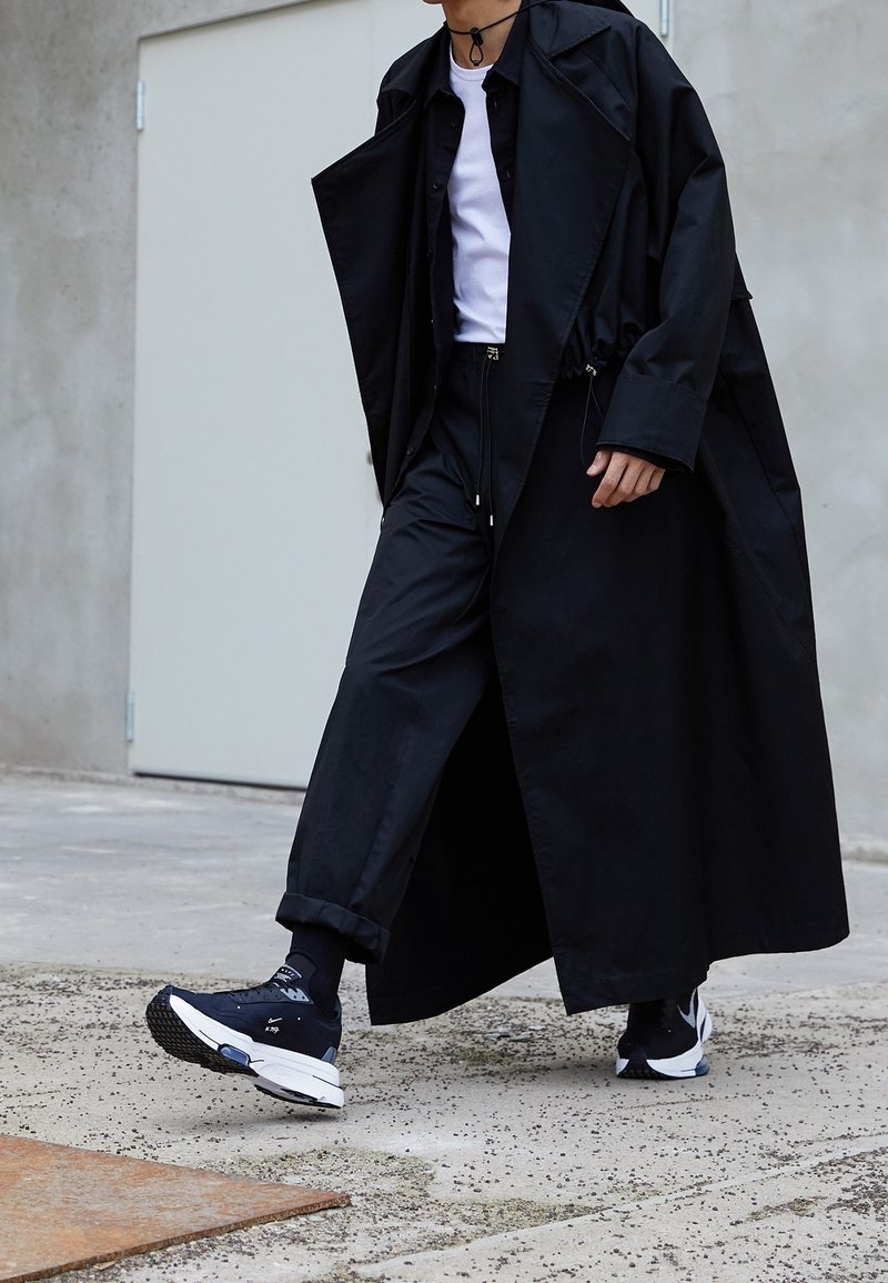 Czarny długi płaszcz na czarnej koszuli i spodniach, w zestawie z czarnymi trampkami i skarpetkami. Płaszcz ma luźny krój i teksturowany materiał.