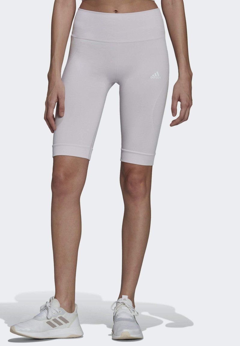 Femme portant des leggings Adidas blancs mi-cuisse et des baskets Adidas blanches, debout les pieds légèrement écartés sur un fond uni.
