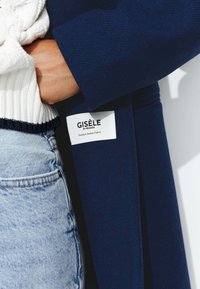 Manteau navy en tissu italien de qualité supérieure, avec une étiquette "GISÈLE par Promod." Associé à un pull blanc texturé et un jean bleu clair.