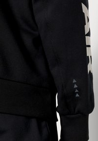 Sudadera negra con puños acanalados, que presenta un gran logo blanco "AIR" en la manga y tres acentos triangulares grises cerca de la muñeca.
