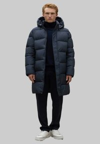 Abrigo acolchado en azul marino oscuro, con capucha, diseño acolchado y un corte hasta la rodilla. Lleva pantalones oscuros y zapatillas blancas.