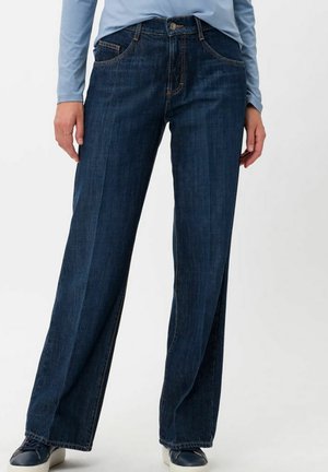 Persona con vaqueros de pierna ancha azul oscuro, camiseta de manga larga azul claro y zapatillas negras, posando contra un fondo blanco liso.