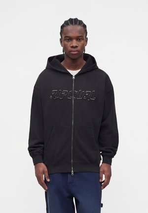 Młody mężczyzna z plecionymi włosami, ubrany w czarną bluzę z kapturem na zamek błyskawiczny z napisem "RIP CURL" i niebieskie spodnie, stoi na białym tle.