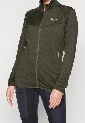 Persoon draagt een olijfgroene jas met ritssluiting en oranje ritszegels en zwarte legging met zijzakken, staand voor een lichte achtergrond.