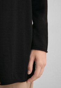 Svart stickad tröja med en mjuk textur, med långa ärmar och rundad fåll. Handen vilar försiktigt vid nederkanten.