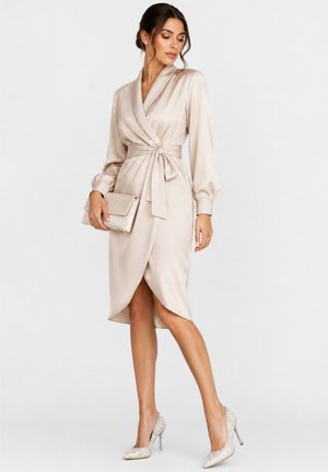 Femme en robe portefeuille en satin beige clair à manches longues, tenant une pochette assortie et portant des chaussures à talons hauts avec des détails en perles.