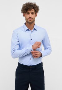 Lichtblauw overhemd met knoopsluiting, kraag, lange mouwen en donkere knopen. Heeft een subtiele textuur en een getailleerd ontwerp. Gecombineerd met donkere broek.