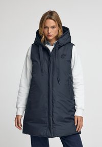 Mörk marinblå pufferväst med stor huva, avtagbara snören och sidofickor. Har en diskret logotyp på bröstet och rak skärning.