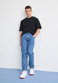 Maglietta nera a maniche corte con logo sul petto, jeans di denim blu chiaro e sneakers bianche con accenti rossi. Outfit semplice e casual.