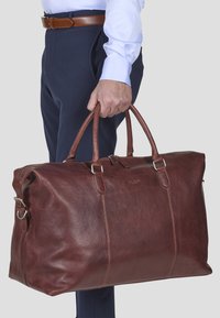 Bruine leren sporttas met twee handvatten, ritssluiting en zilveren hardware, met een gladde textuur en minimalistisch design.