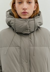 Grijze donsjas met een hoge kraag, voorzien van drukknopen, een trekkoordsluiting en een doorgestikte textuur. Gladde stof met een matte afwerking.