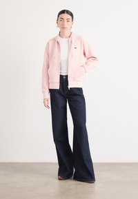 Sweat à capuche rose zippé avec un logo, porté sur un T-shirt blanc. Associé à un jean en denim sombre à jambe large ; texture lisse, design décontracté.