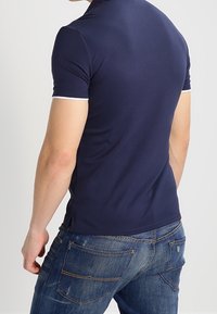 Mörkblå piké med korta ärmar som har vita accenter på kragen och ärmarna, tillverkad av mjukt, elastiskt tyg. Bärs tillsammans med denimjeans.