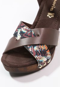 Sandale compensée marron avec des brides croisées, une en cuir uni et l'autre en tissu multicolore à motifs avec des clous métalliques, et une semelle intérieure rembourrée.