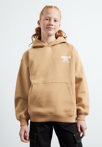 Felpa beige con tasca frontale, polsini a coste e logo bianco. Abbinata a pantaloni cargo neri con tasche laterali.