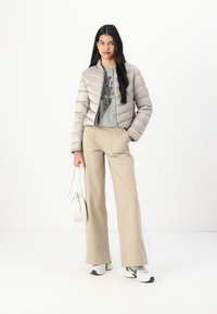 Light gray puffer jacket, zippered, with horizontal padding. Grey graphic t-shirt underneath, paired with beige wide-leg trousers and sneakers.