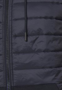 Giacca a vento navy imbottita con pannelli cuciti, zip frontale e cappuccio con cordino e fermaglio nero. Texture morbida e liscia.