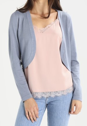 Femme portant un cardigan court bleu clair sur un caraco rose pâle à dentelle, associé à un jean bleu clair sur fond blanc.
