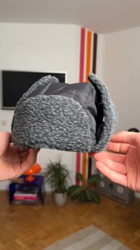 Šedý klobouk s plyšovým, texturovaným povrchem a hladkým vnitřním podšívkou. Má zaoblený tvar a měkké náušníky pro větší teplo.