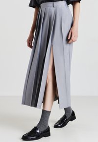 Jupe grise plissée avec fentes avant, design ajusté, texture lisse, associée à des chaussettes grises mi-mollet et des mocassins noirs.