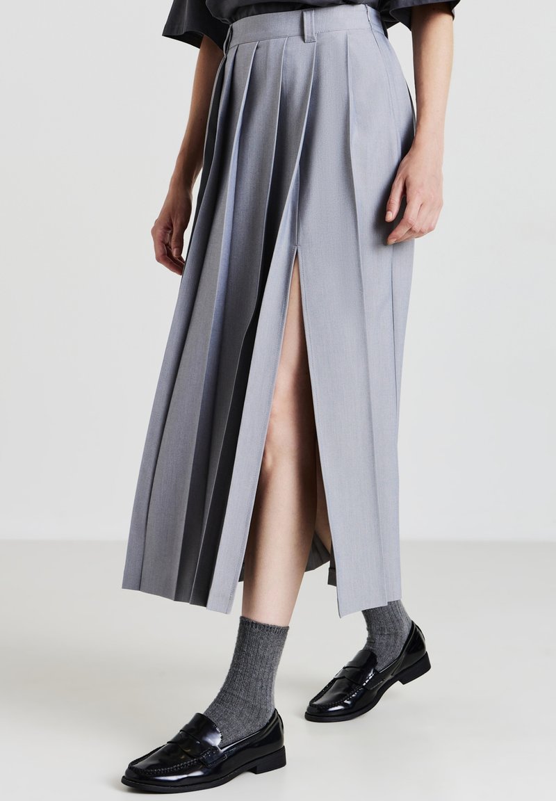 Jupe grise plissée avec fentes avant, design ajusté, texture lisse, associée à des chaussettes grises mi-mollet et des mocassins noirs.