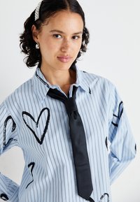 Blårandig skjorta med svarta hjärtformer, med en klassisk krage och ett svart slips. Slät tyg med en avslappnad passform.