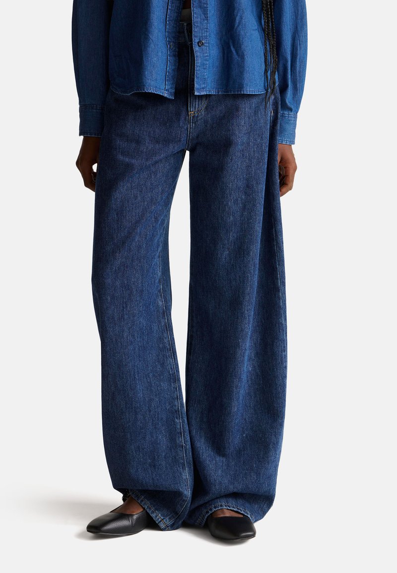 Vaqueros de pierna ancha en azul oscuro, fabricados en algodón, con un diseño clásico de cinco bolsillos y un ajuste relajado. Combinados con zapatos planos negros.