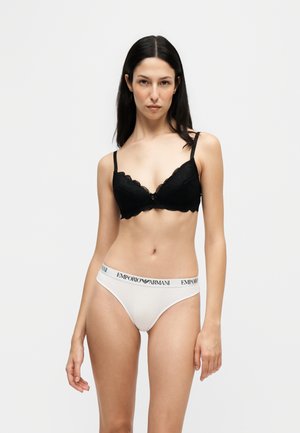 Femme aux longs cheveux foncés portant un soutien-gorge en dentelle noire et un slip blanc avec le logo Emporio Armani sur un fond clair uni.