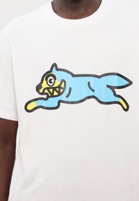 Hvid t-shirt med et blåt tegneseriehundedesign med et gult ansigt og sorte konturer, der præsenterer en legesyg pose og abstrakte træk.